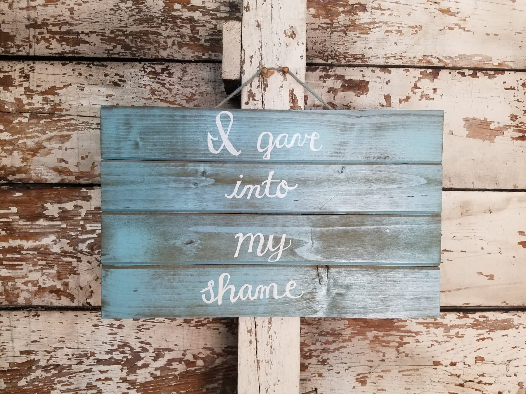 blue faux-weathered sign with "I gave into my shame" painted on it in white script
