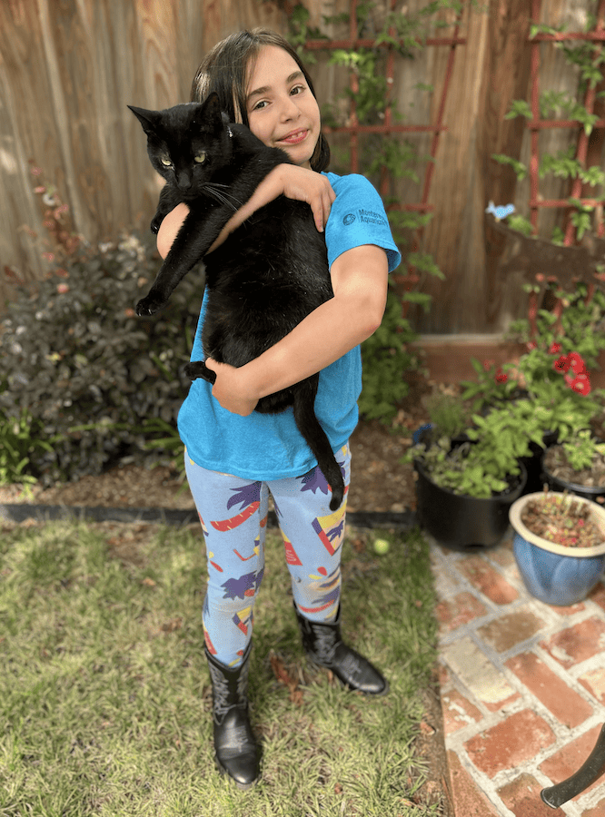 Snarly's niece Lucy holds her black cat, Alex, in her backyard, wearing a blue t-shirt and blue palm-tree-patterned Nooworks leggings