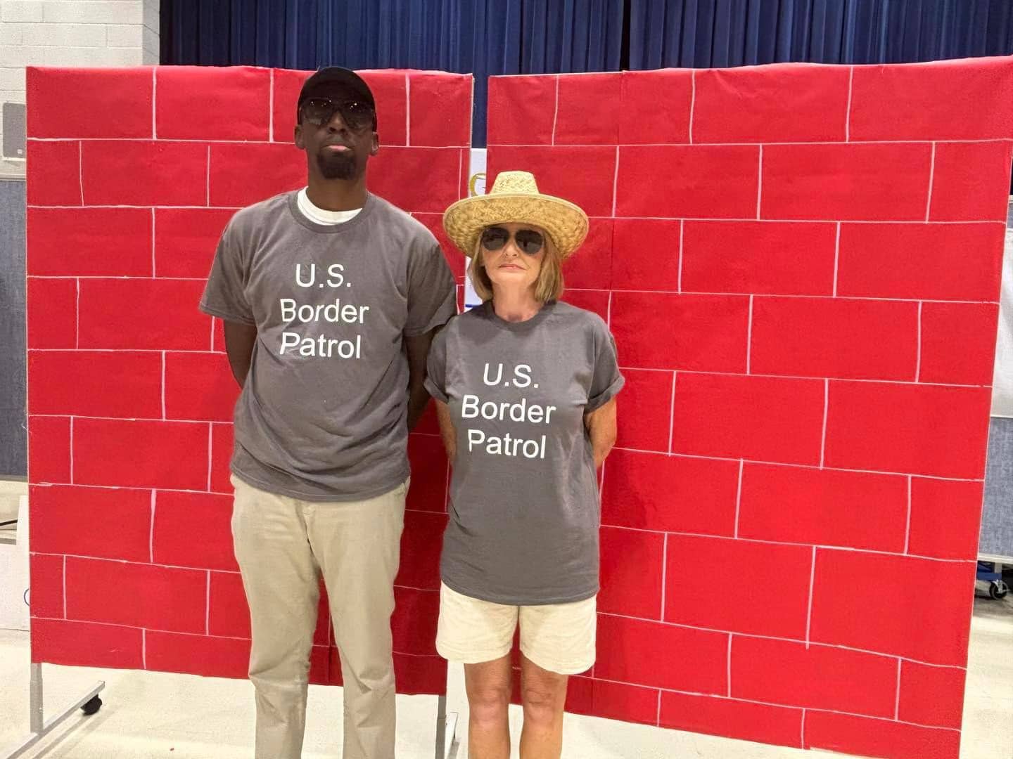 two adults wearing gray t-shirts reading "U.S. Border Patrol," standing in front of a giant bright-red "brick" wall. 