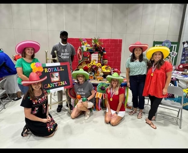 Image depicts adults in bright sombreros in front of a sign that says Royall Cantina, plus a large brick wall; two adults are wearing gray shirts that read "Border Patrol." 