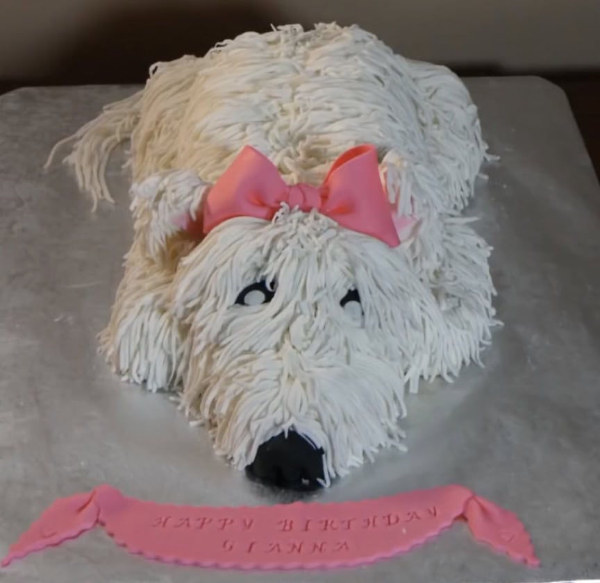 Cake carved and frosted to look like a recumbent shaggy dog wearing a pink bow.